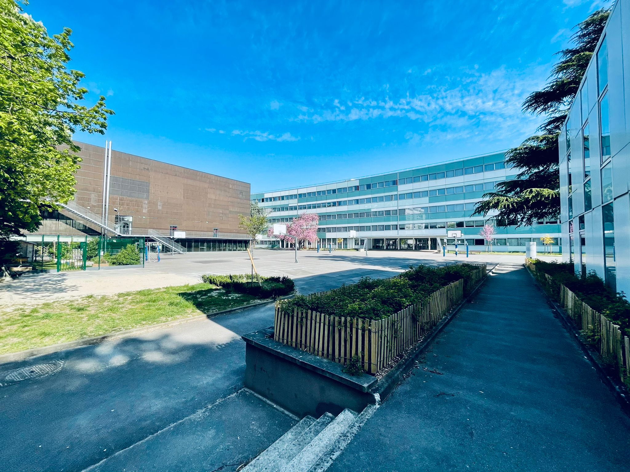 Cour, avec les terrains de sport, des petites bordures d'espace vert, le gymnase à gauche et le lycée au fond