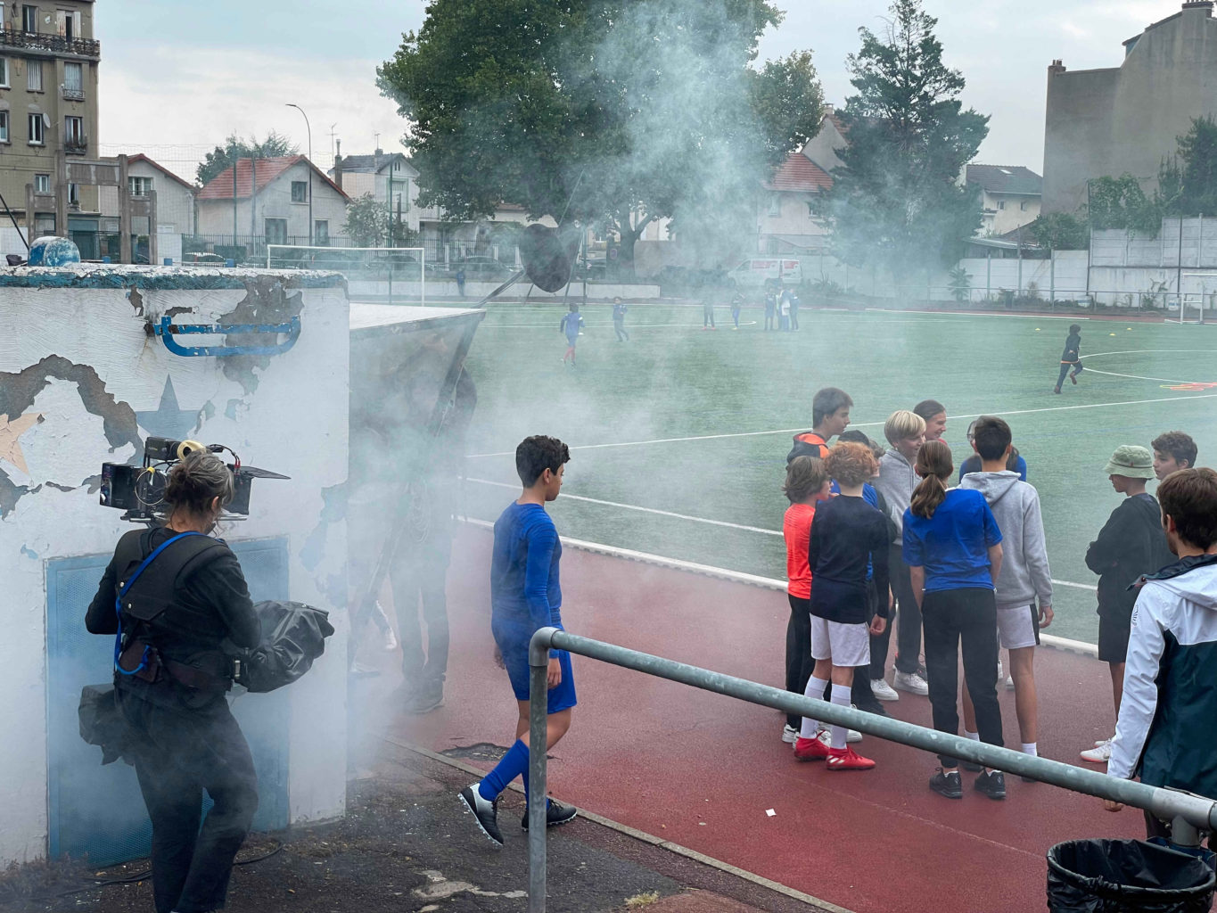 Sur un terrain de sport (extérieur) et dans un nuage de fumée artificielle, la caméraman filme les élèves