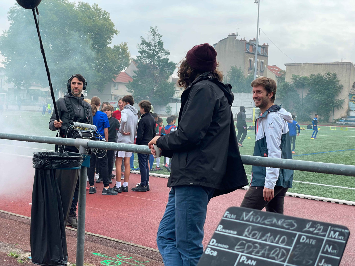 Sur un terrain de sport (extérieur) des membres de l'équipe technique au premier plan avec les élèves (dans un nuage de fumée artificielle) au second plan